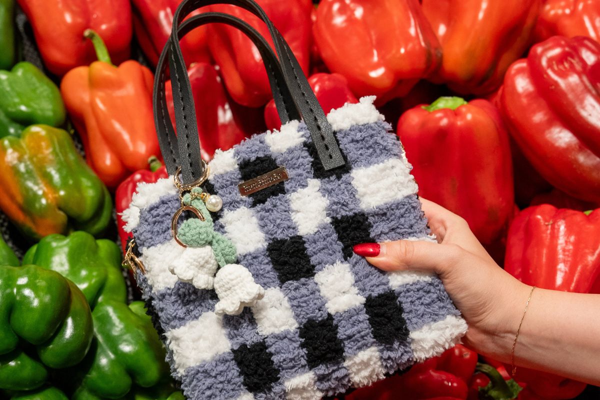 Hand holding a handmade woven handbag with a fluffy black-and-white checkerboard design, black handles, and a crochet flower charm, set against a vibrant background of red and green capsicums.