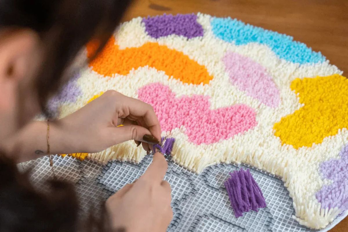 Close-up of hands using a latch hook tool to attach colorful yarn onto a canvas, demonstrating the rug-making process.