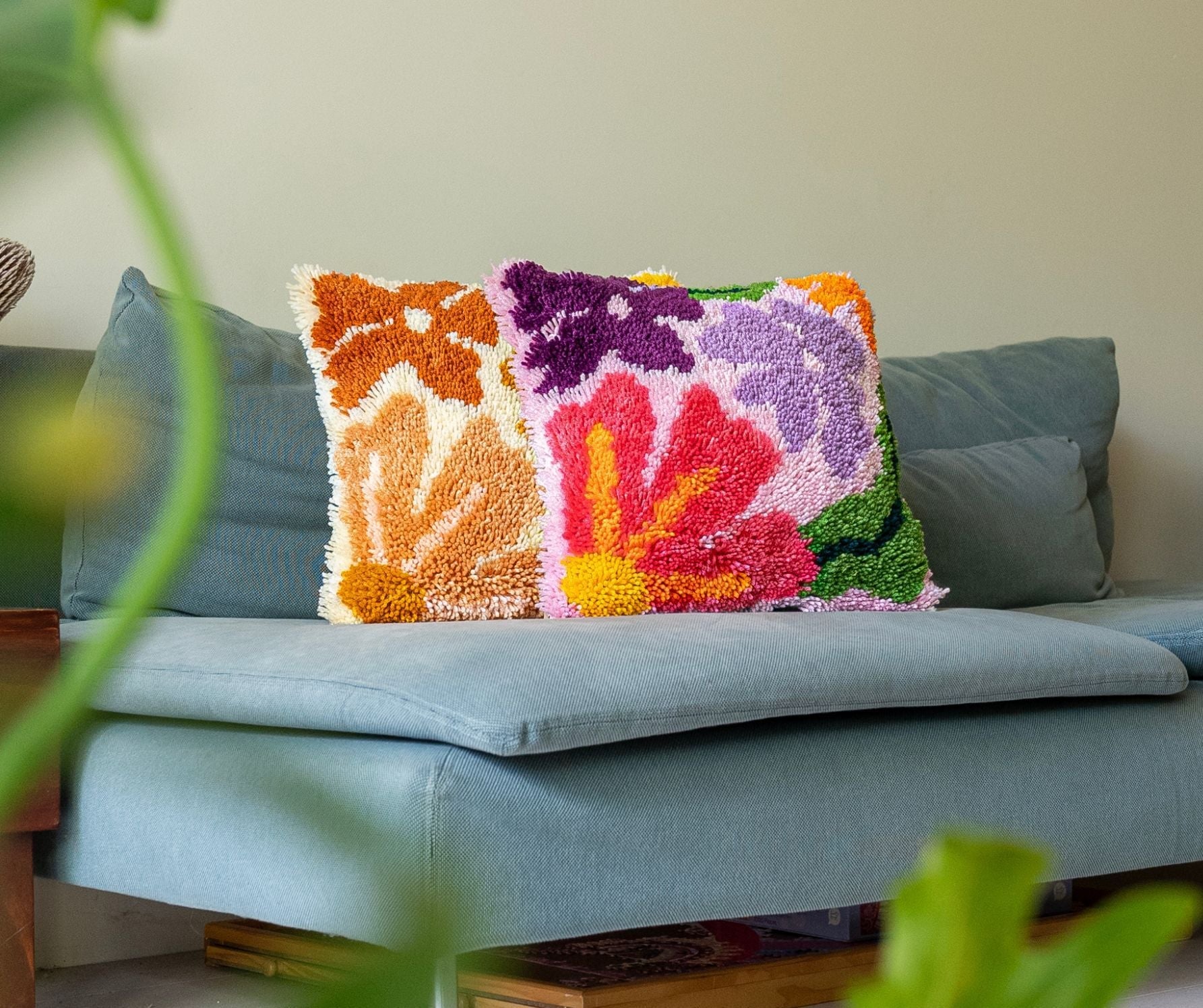 Three latch-hooked cushions sit on a white couch, a hand caresses one of the cushions. The cushion on the left is brown, the one in the middle is purple and the one on the right is green & pink