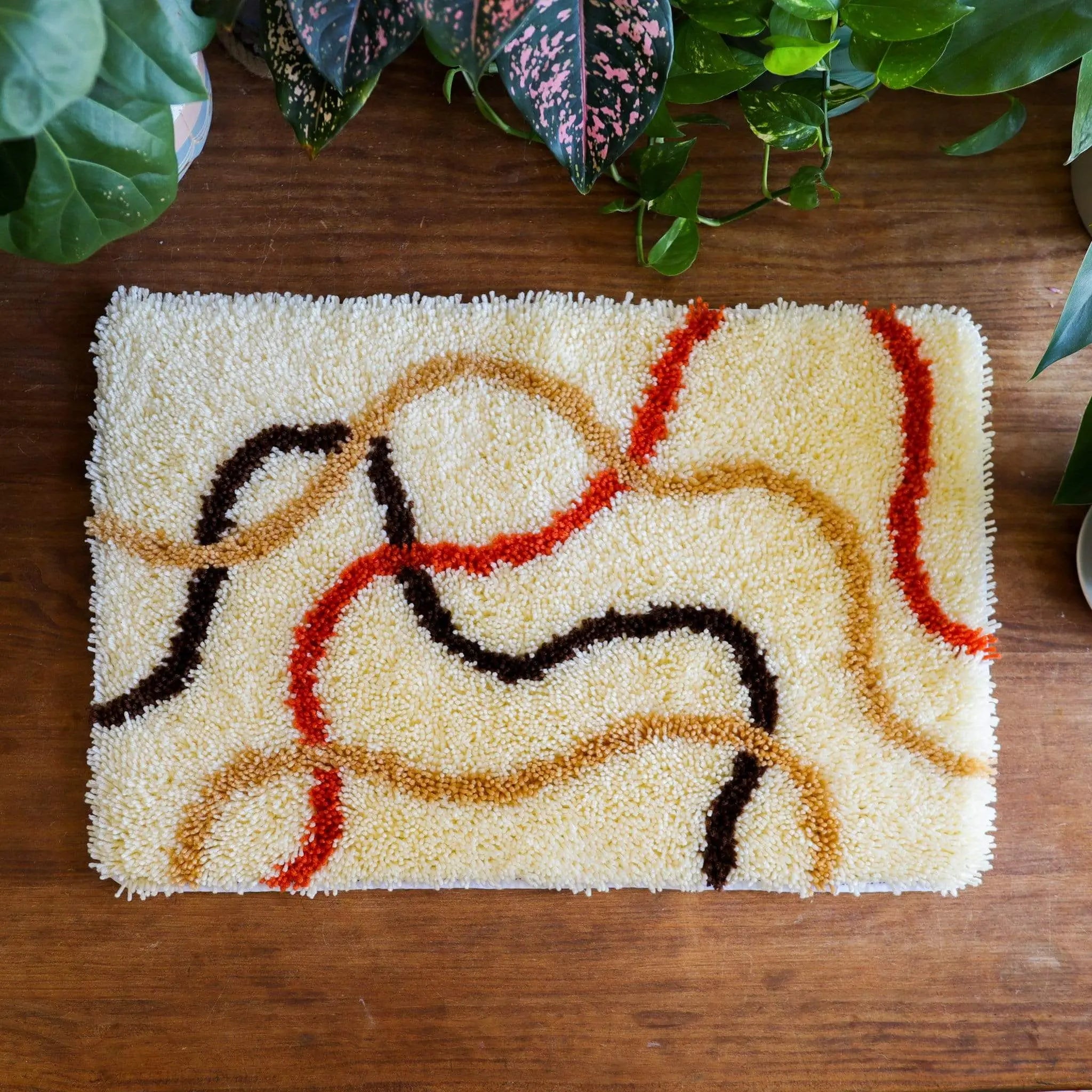 Craft Club Co FLOW Rug Making Kit. This rug is a rectangle shape and has a cream background with swirling lines across the design. the lines are in light brown, dark brown and deep orange.