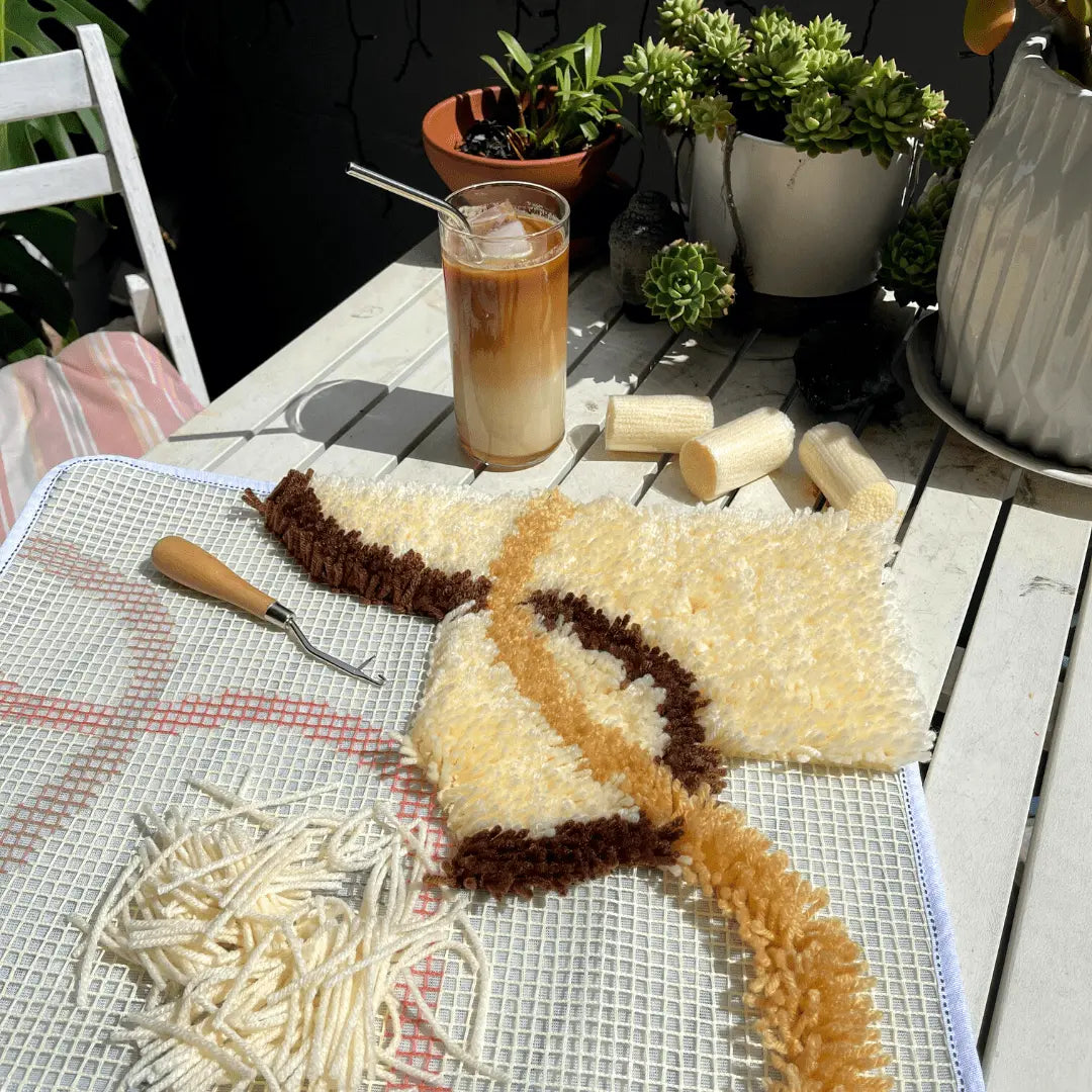 Craft Club Co FLOW Rug Making Kit. An incomplete rug is shown on an outdoor table with pot plants and an iced coffee. There are cream pieces of yarn and a latch-hook sitting on the rug backing.