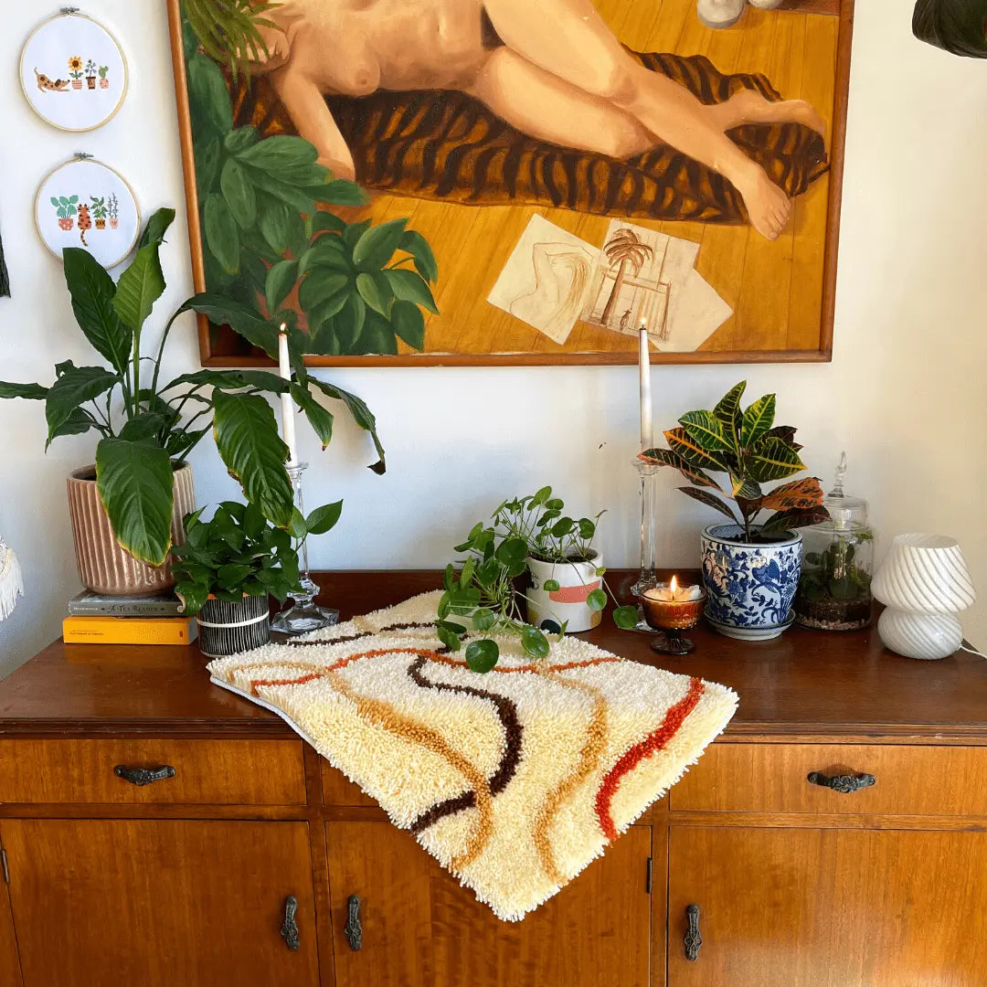 Craft Club Co FLOW Rug Making Kit. A midcentury modern mantel piece is shown with the rug placed on it as decor. The mantel piece also has pot plants and candles on it, with a large oil painting behind it.